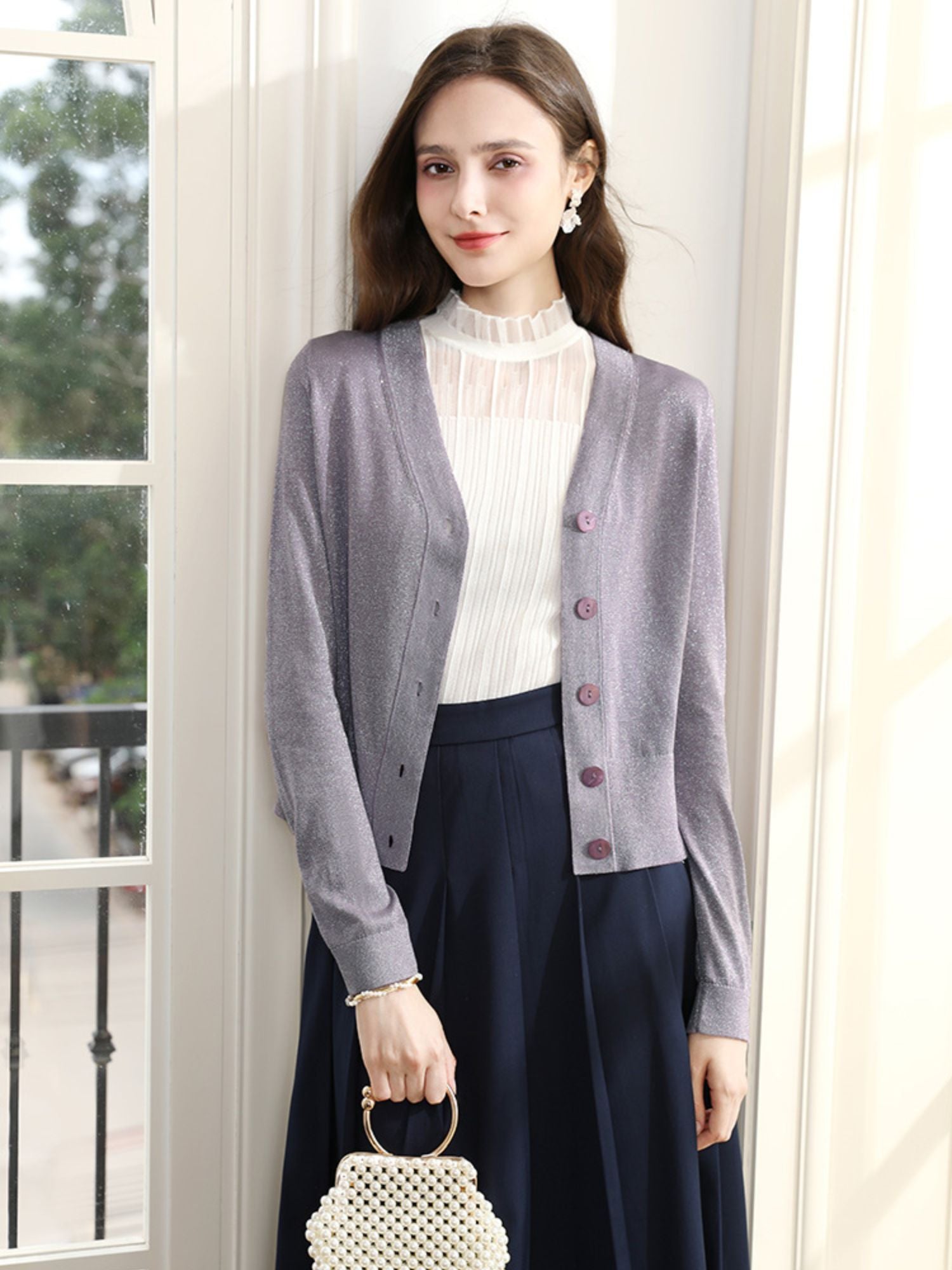 Woman in lavender cardigan and cream blouse posing indoors with a neutral expression.