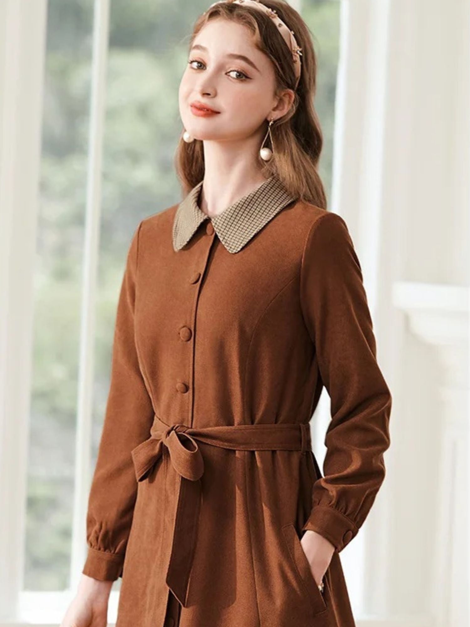 Woman posing in brown dress, holding her collar and standing near a window.