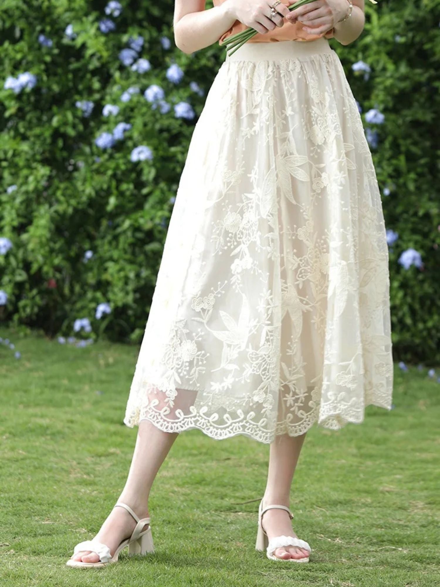 Woman in peach t-shirt and ivory lace skirt standing on grass with legs slightly crossed.