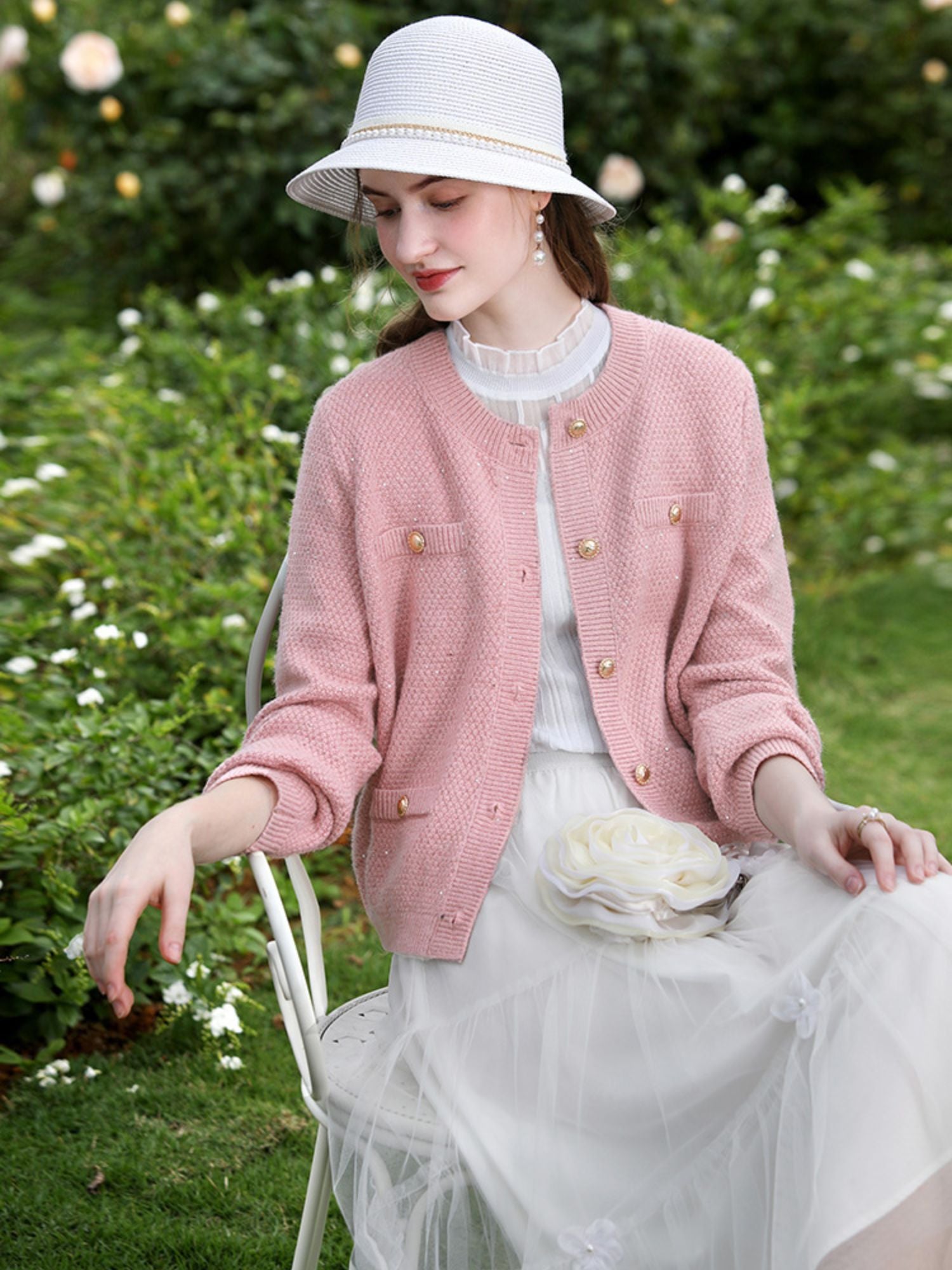 Woman in pink jacket and tulle skirt with a white hat, touching a flower gently.