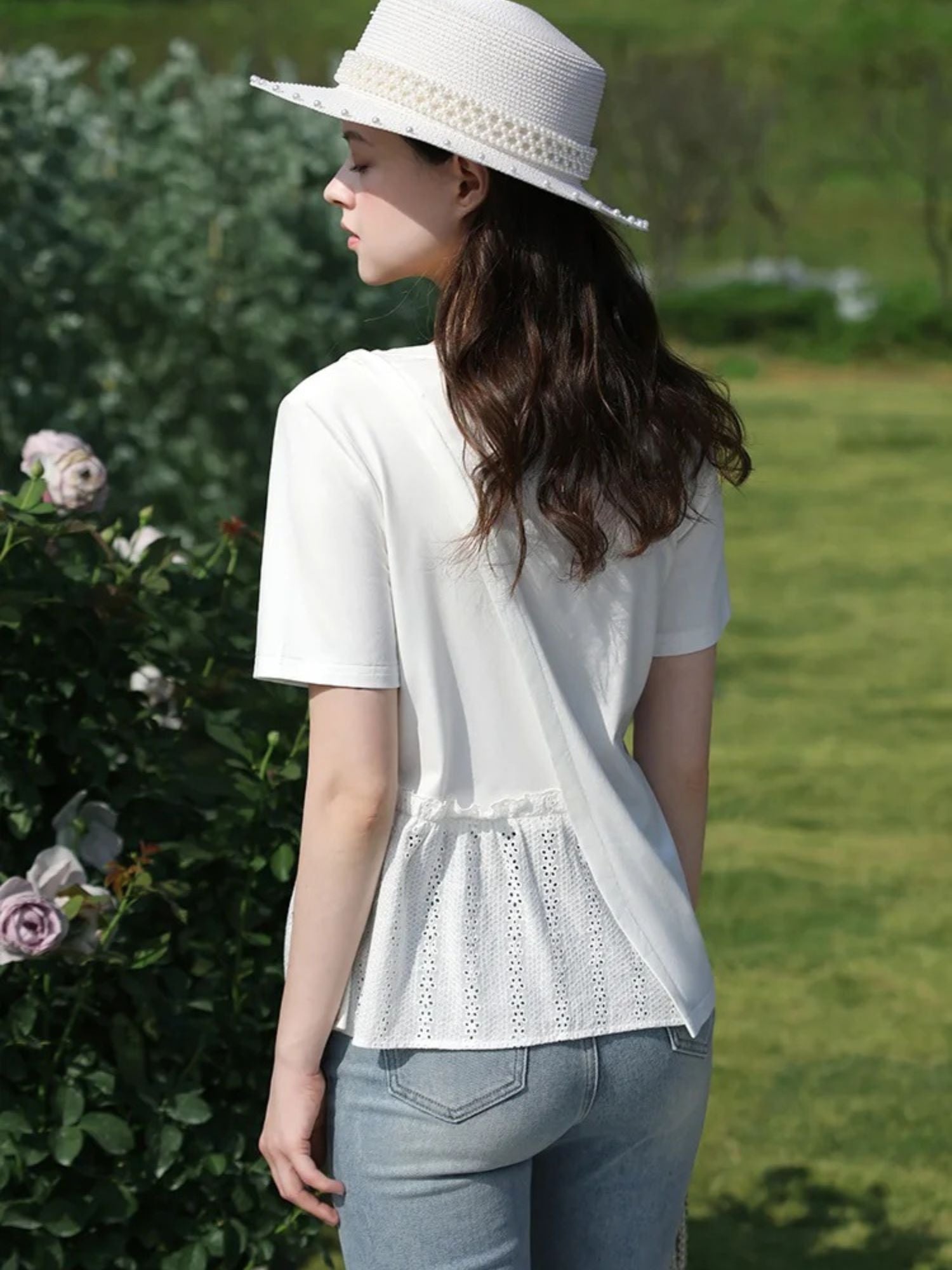 Back view of woman in lace-trimmed blouse and hat, walking beside flowering bushes.