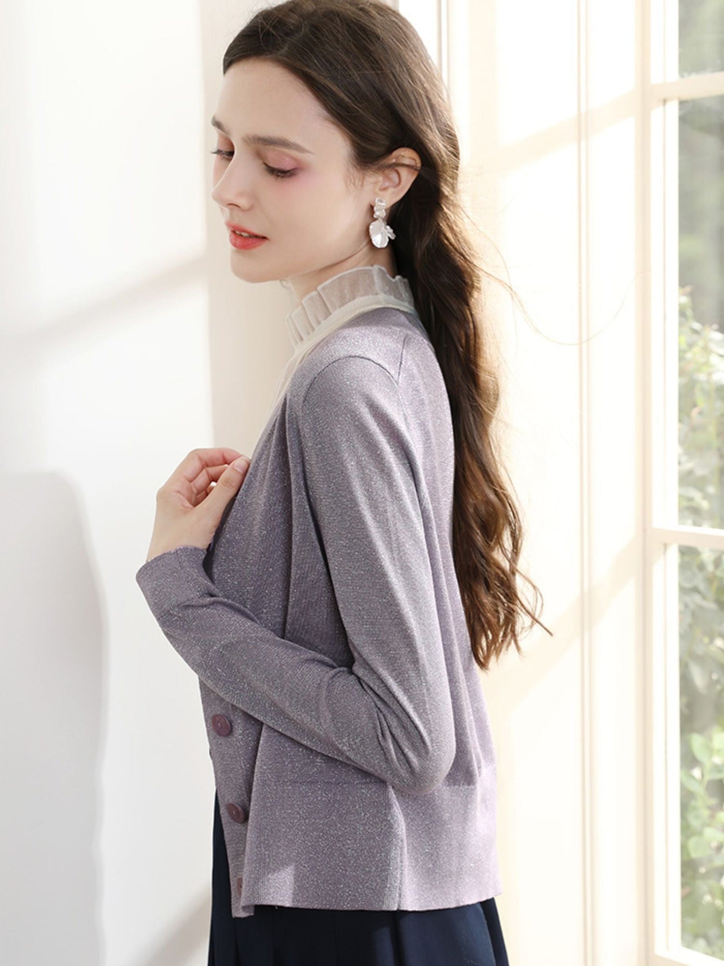 Woman standing sideways in purple cardigan, gently holding the strap of her handbag.