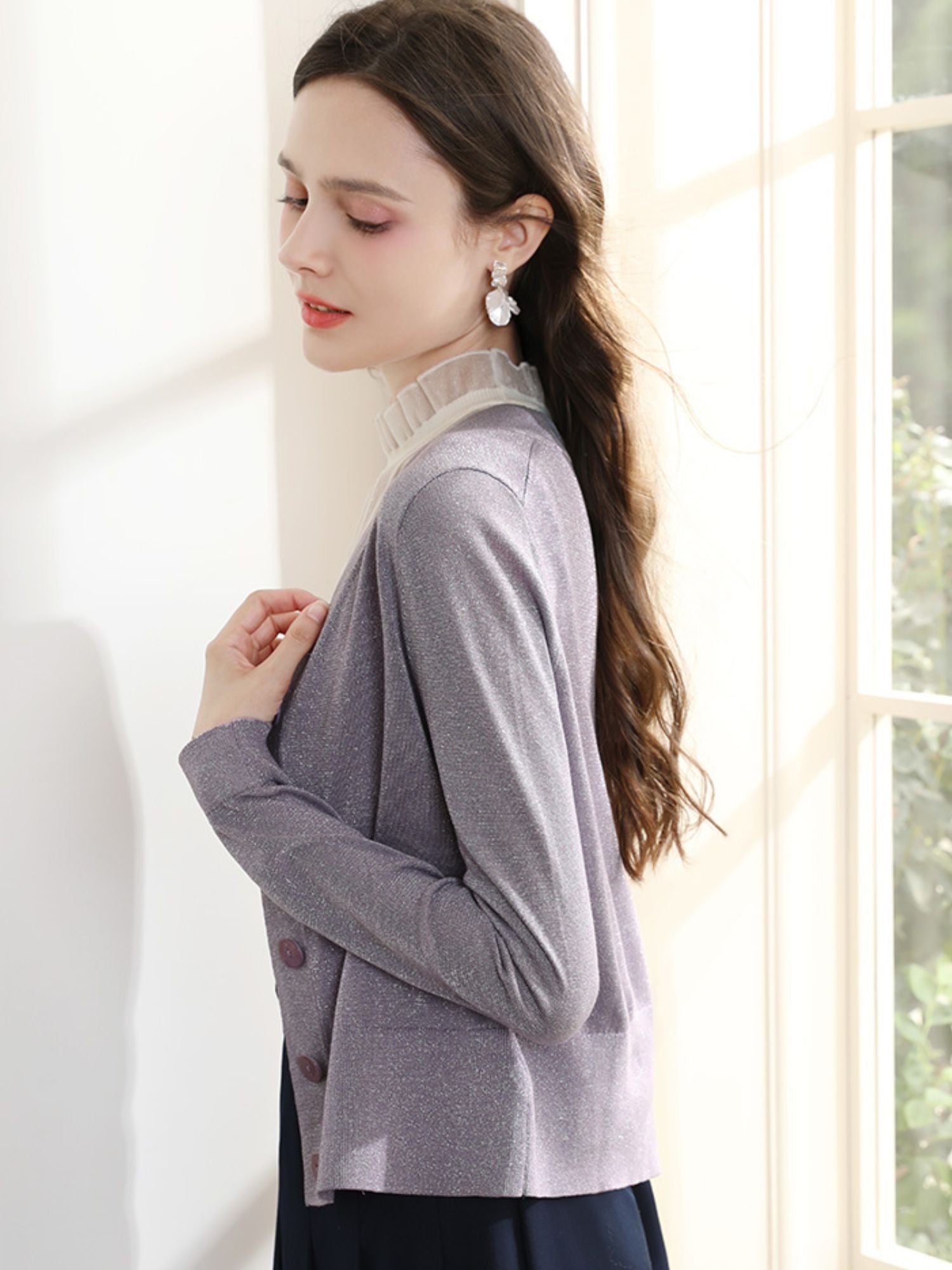 Woman standing sideways in purple cardigan, gently holding the strap of her handbag.
