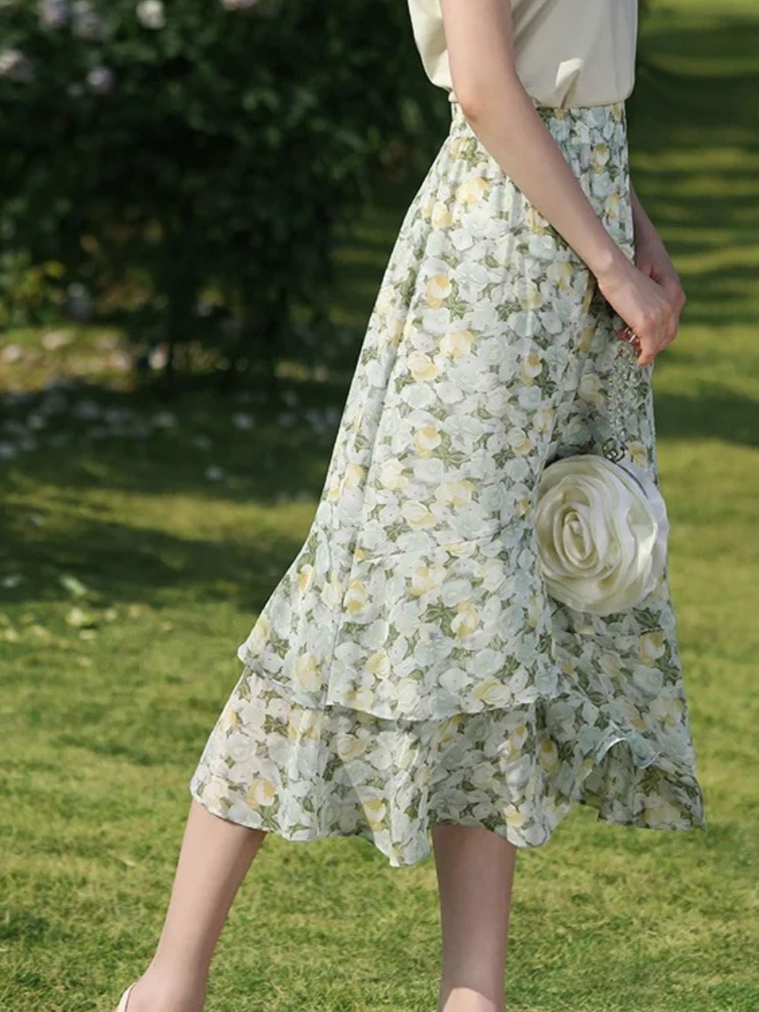 Woman holding a small round bag while wearing a floral skirt and pale blouse on the grass.