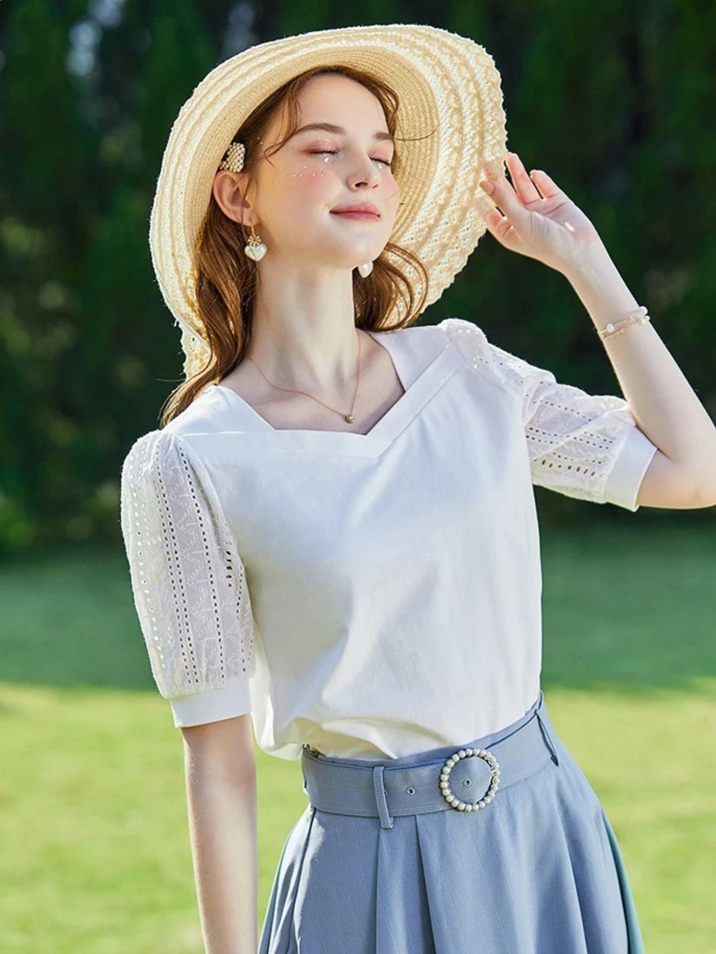 Woman in white lace-sleeve top and blue skirt, smiling under a straw hat.
