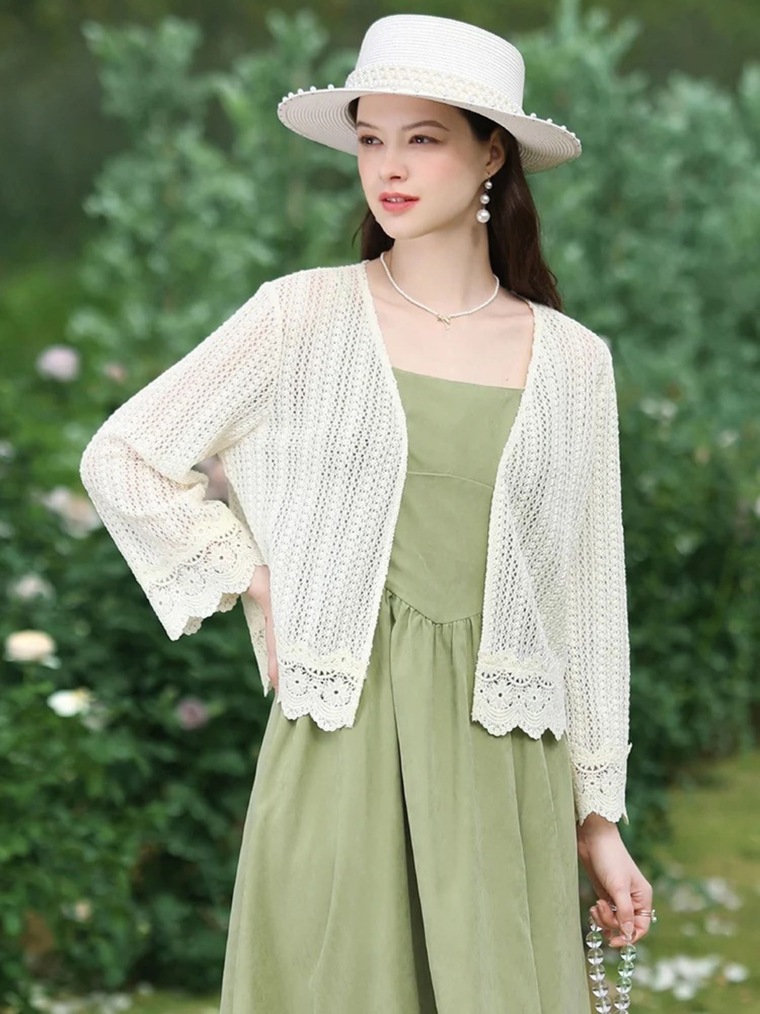 Woman lifting her arm slightly in a white lace cardigan and green dress, smiling outdoors.