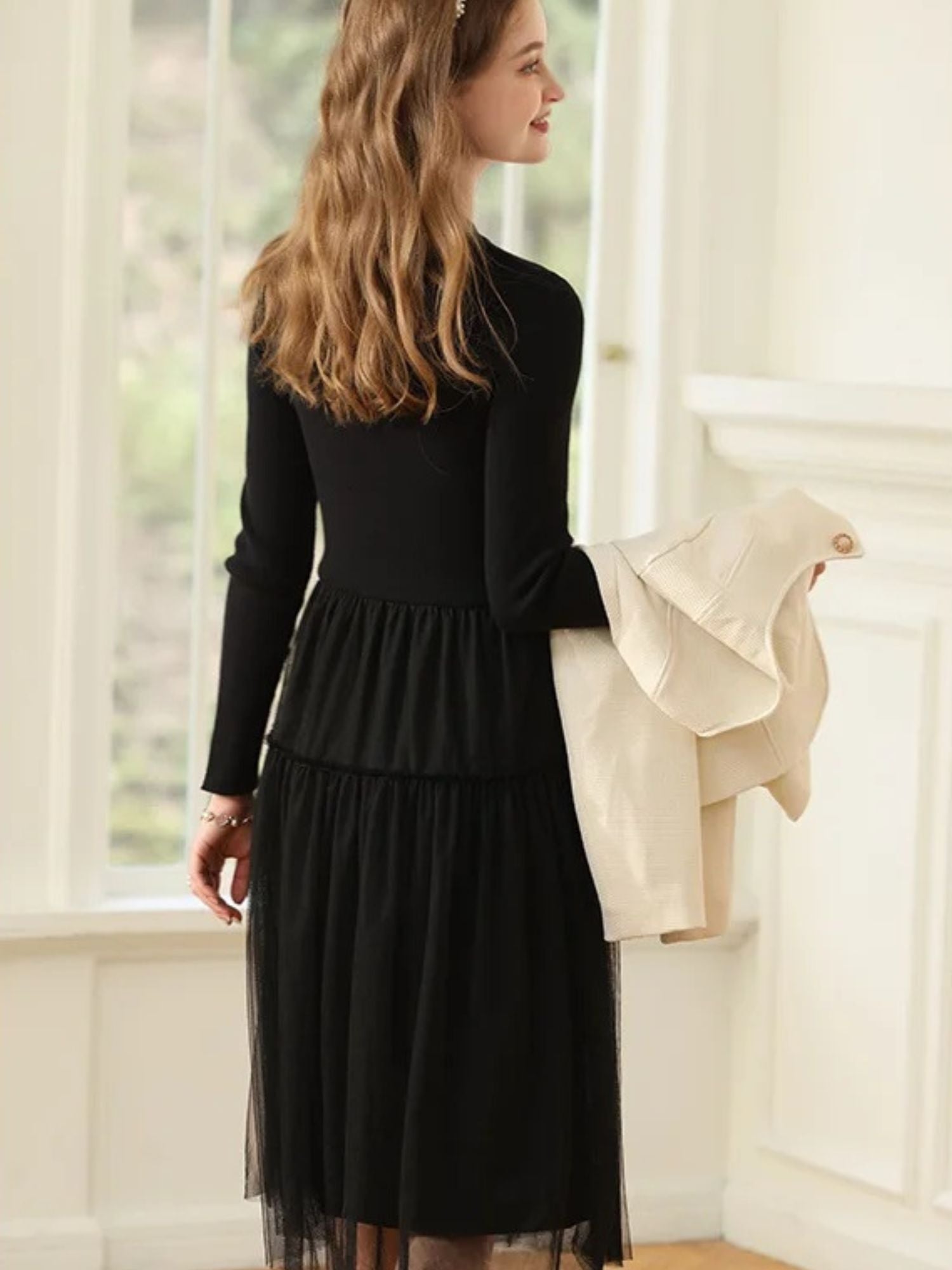 Woman smiling in black dress, holding a light coat with one hand on her hip.