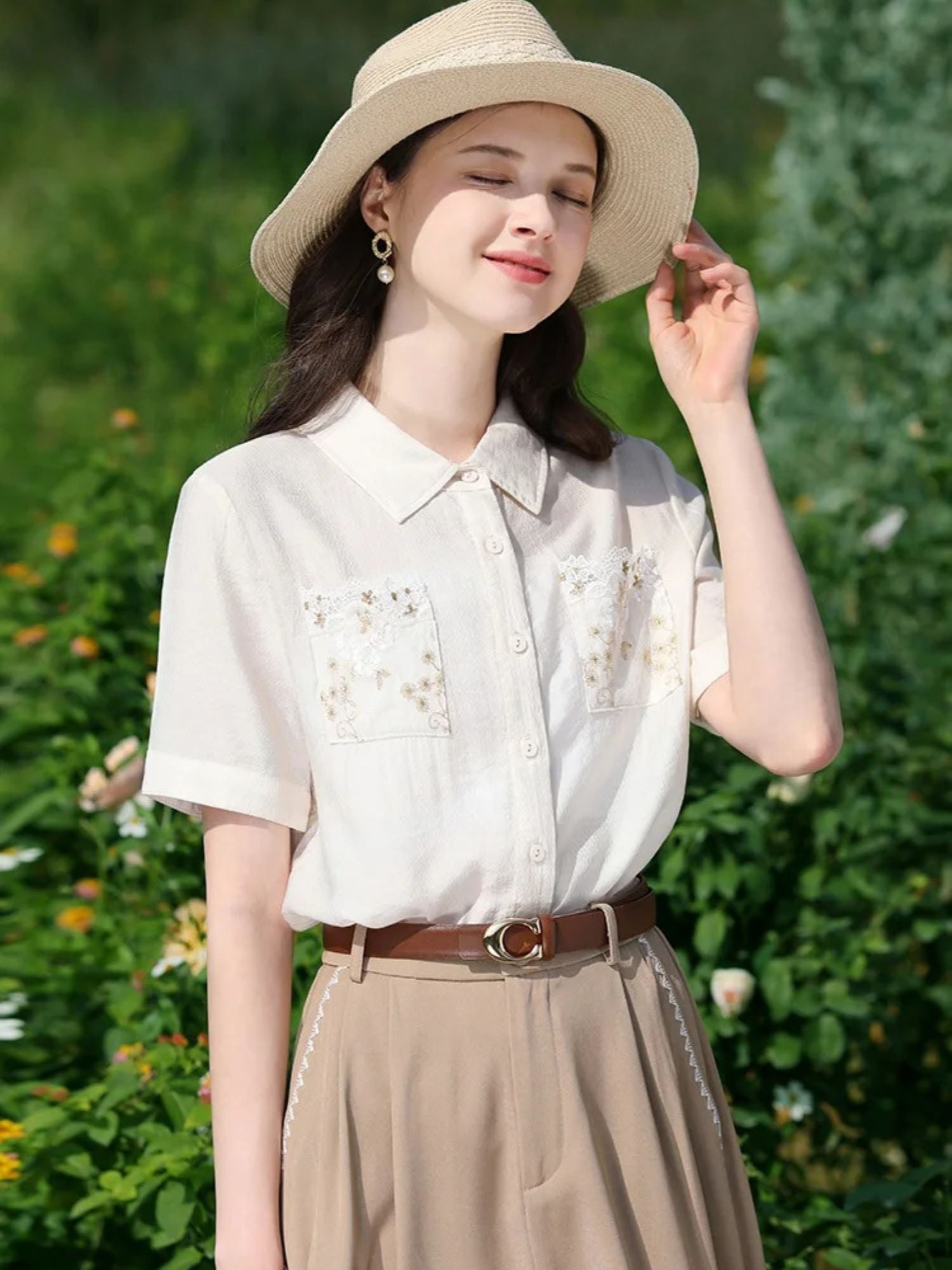 Woman in floral blouse and flowy skirt smiling while holding her hat.