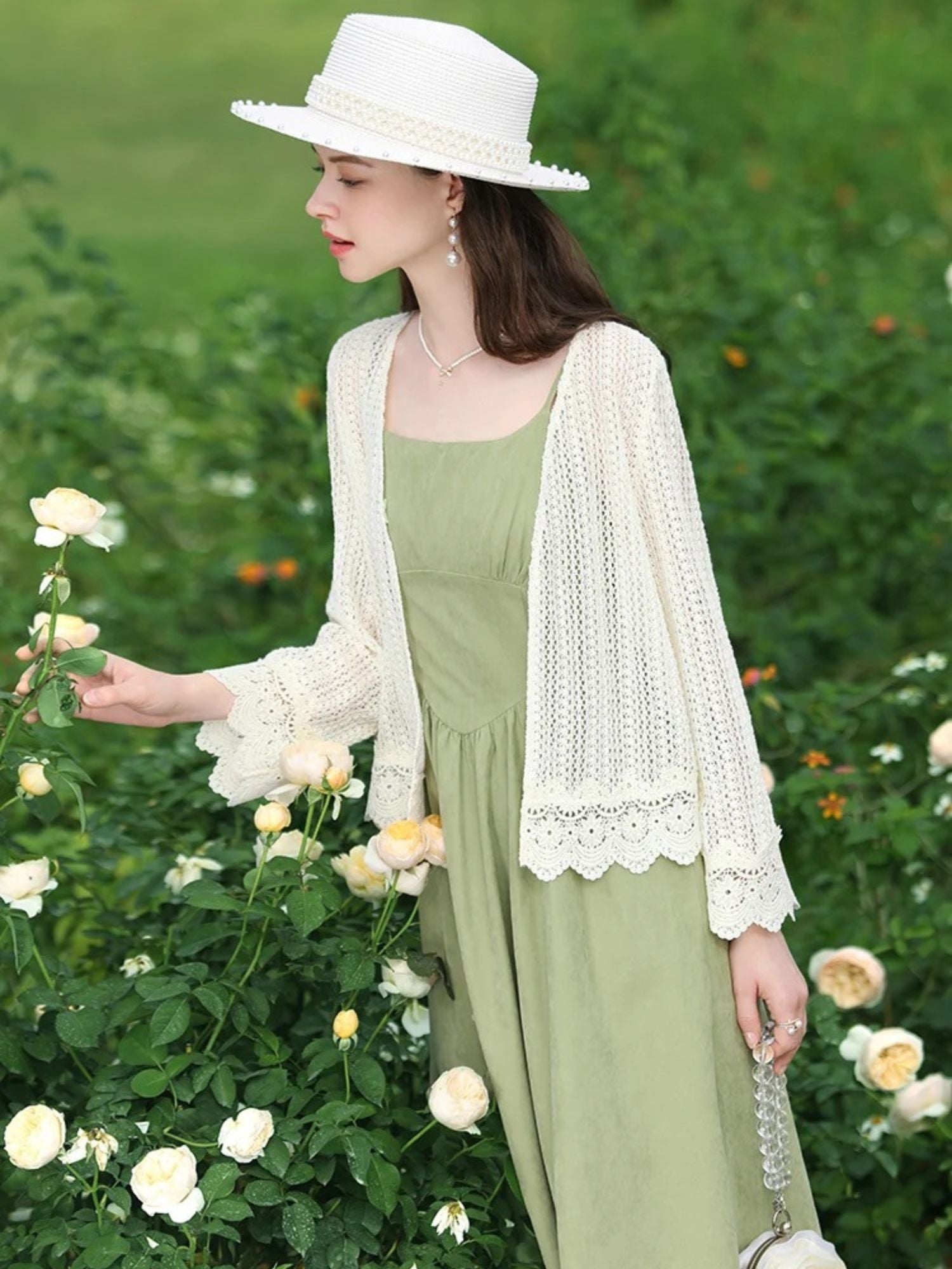 Woman in lace cardigan holding a flower, walking in a rose garden with a soft expression.