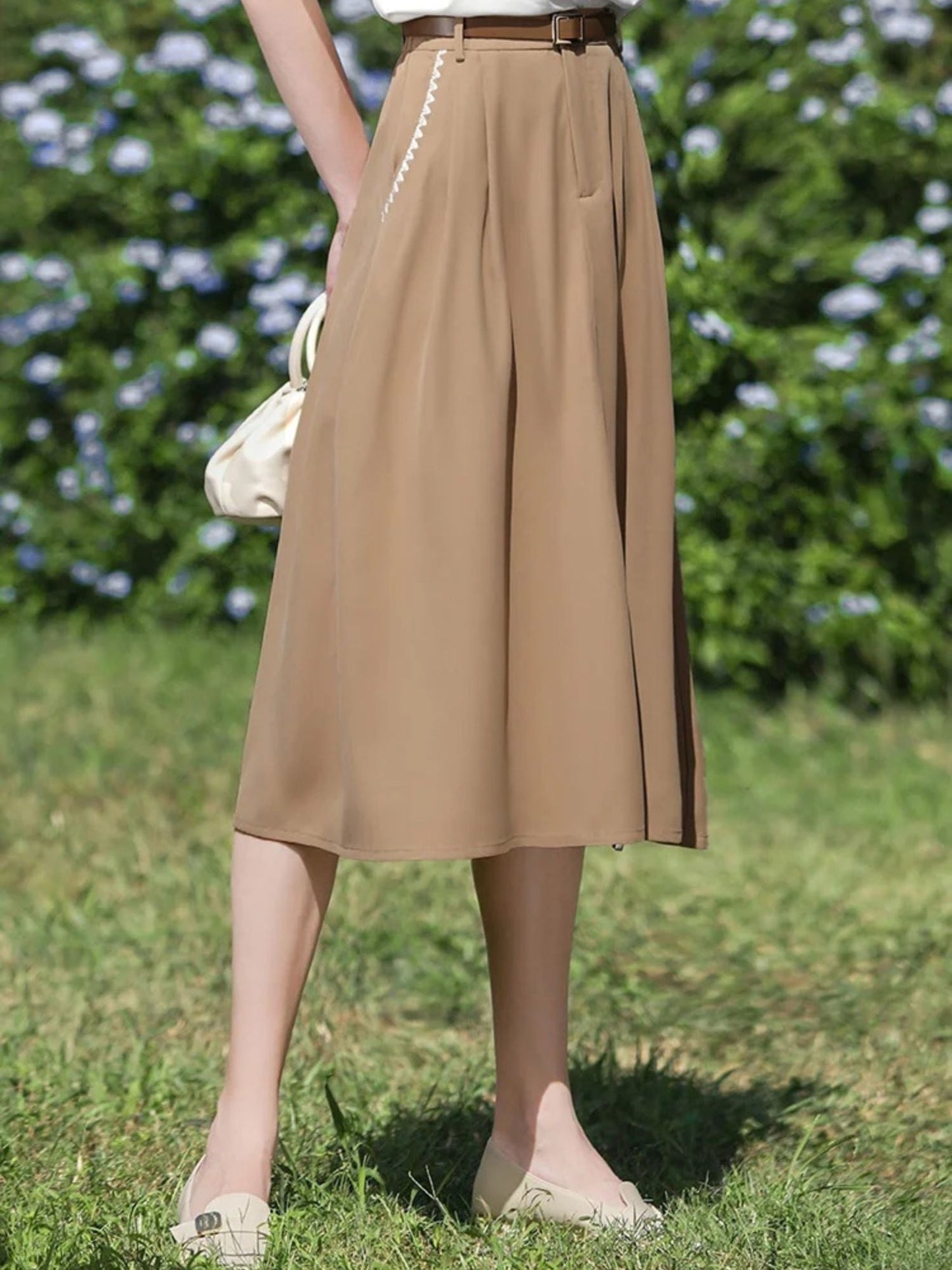Woman wearing tan skirt and white tee with one leg slightly bent while standing on grass.