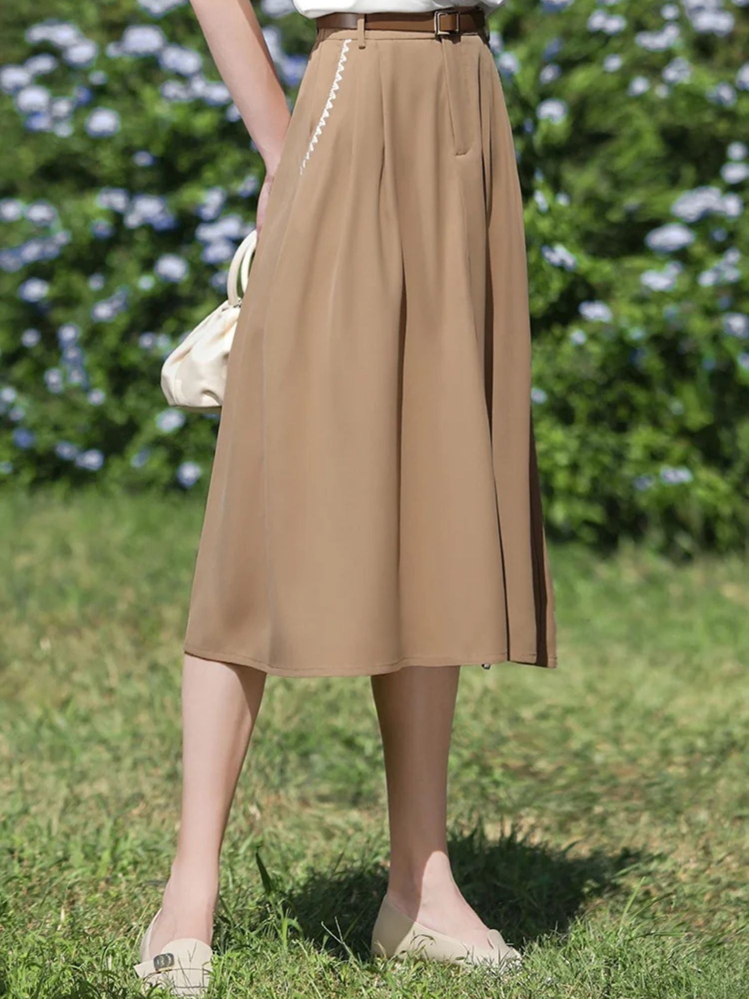 Woman wearing tan skirt and white tee with one leg slightly bent while standing on grass.