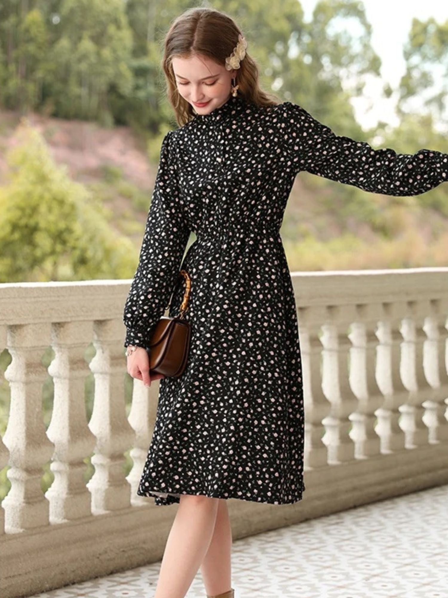 Woman in black floral dress walking on a balcony, holding a small brown purse.