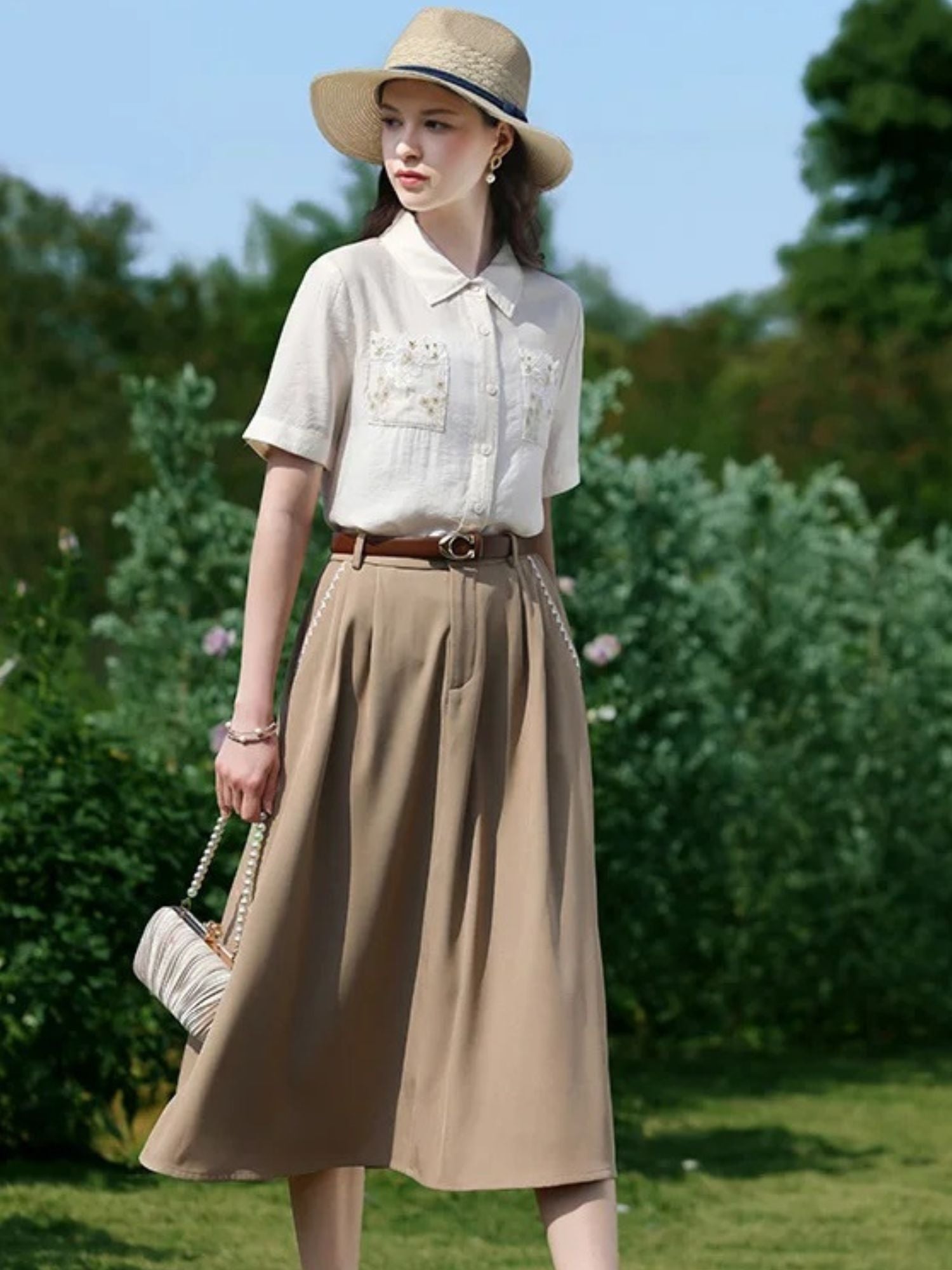 Woman in embroidered blouse and belted skirt walking on grass with a woven handbag.