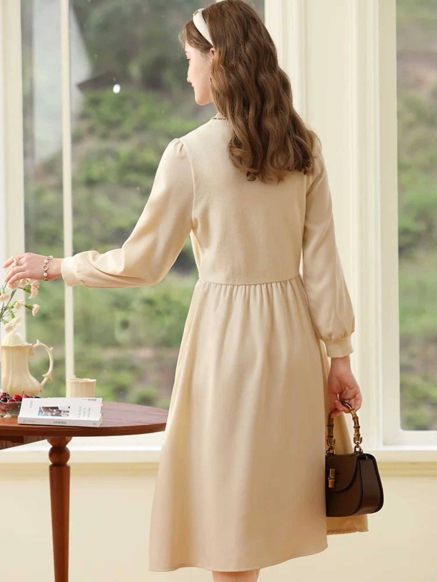 Back view of woman in cream dress walking toward a window, holding a handbag.