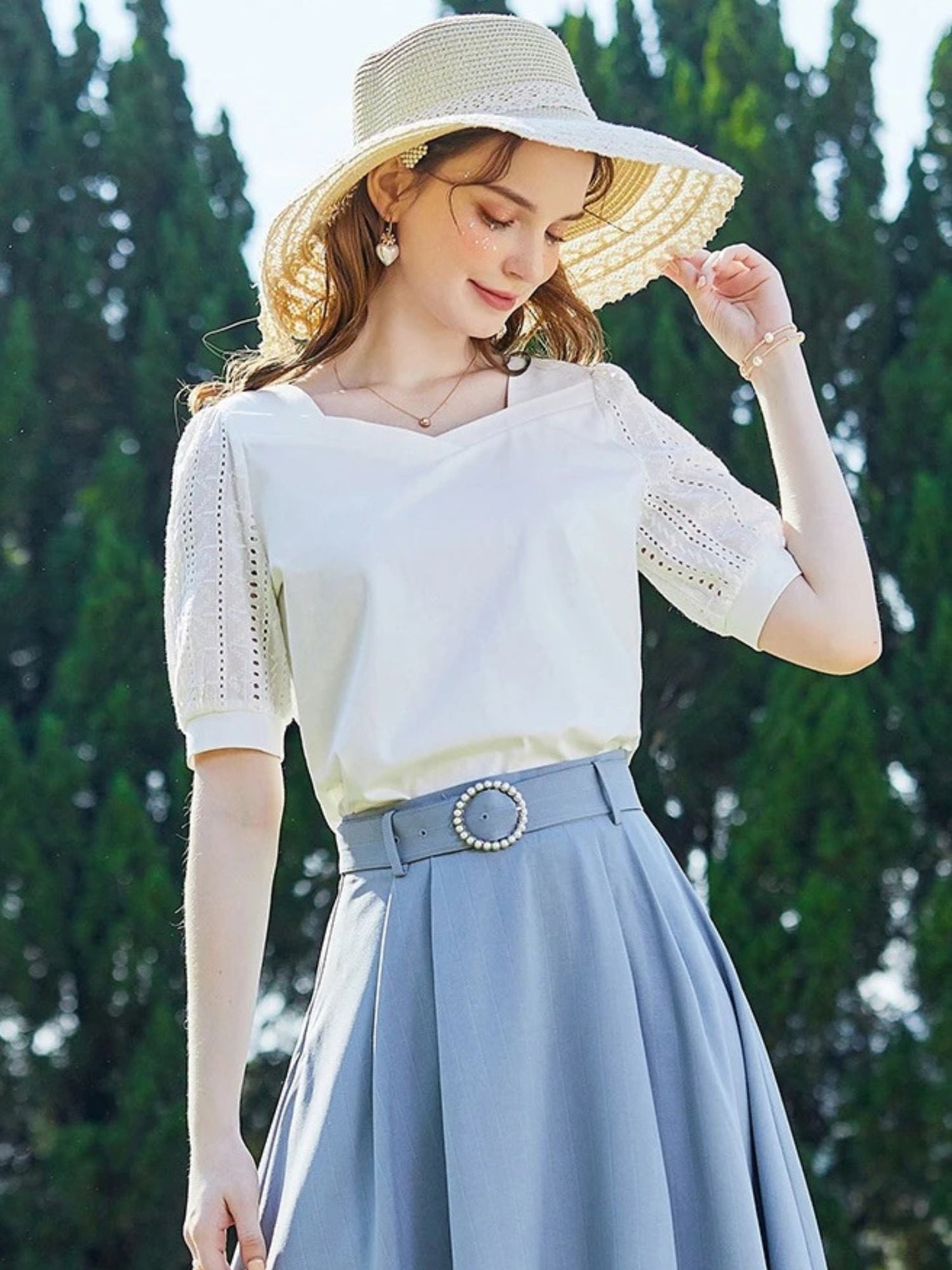 Woman in white lace top and blue skirt, adjusting her hat near bushes.